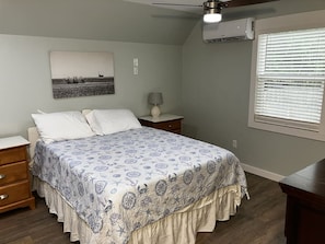 Un dormitorio con cama, mesitas de noche, lámpara, ventilador de techo y ventana con persianas.