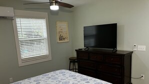 Une chambre à coucher comprenant un lit, une commode, un téléviseur et une fenêtre avec des stores.
