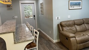 Un salon avec un canapé en cuir marron, un coin repas doté d’un comptoir en granit et de chaises blanches, et une porte donnant sur un balcon.