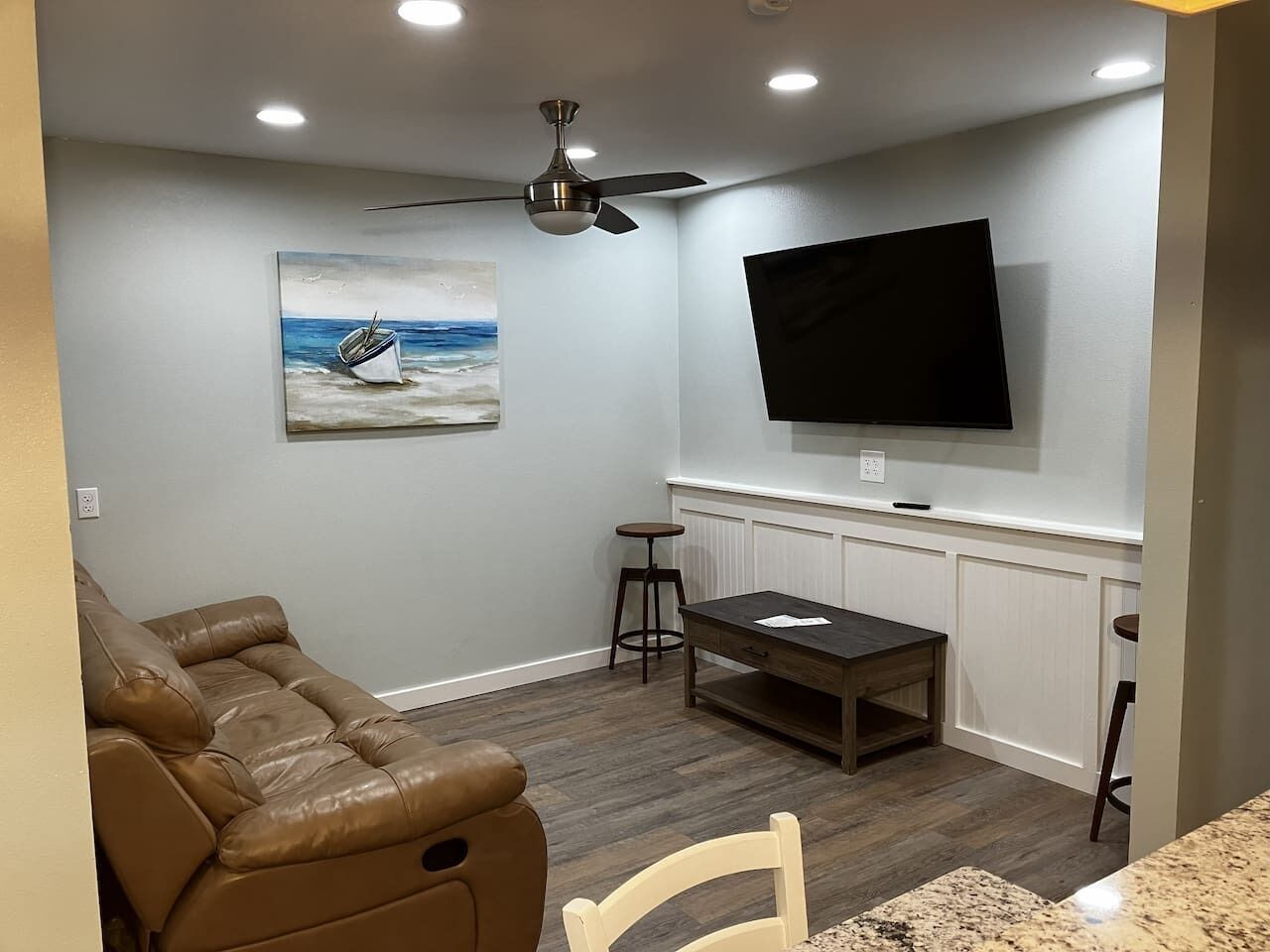Een woonkamer met een bruine leren bank, een eettafel met twee stoelen, een plafondventilator, een flatscreen-tv aan de muur en een schilderij van een boot.