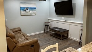 Un salon avec un canapé en cuir marron, une table à manger avec deux chaises, un ventilateur de plafond, un téléviseur à écran plat fixé au mur et un tableau représentant un bateau, également accroché au mur.