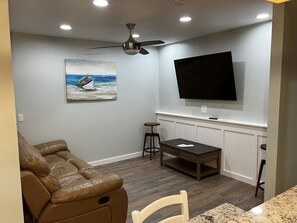 A living room with a brown leather sofa, a dining table with two chairs, a ceiling fan, a flat-screen TV mounted on the wall, and a painting of a boat on the wall.