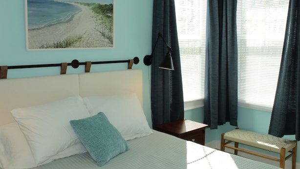 Une chambre à coucher avec un lit, des oreillers, une table de chevet, un banc, un tableau et une fenêtre avec des rideaux.