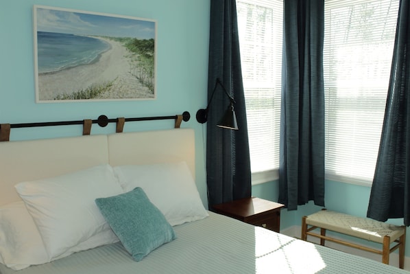 A bedroom with a bed, pillows, a bedside table, a bench, a painting, and a window with curtains.