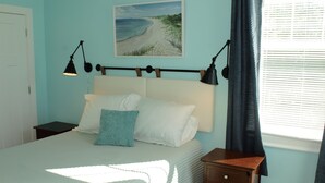 Quarto com cama, mesas de cabeceira, um criado-mudo, janela com persianas e um quadro na parede.