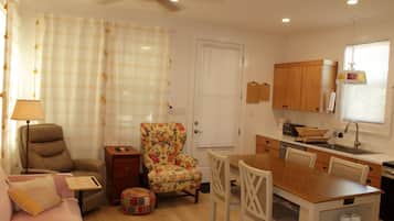 Uma sala de estar acolhedora com sofá, poltrona e uma pequena mesa. Há uma área de cozinha com armários de madeira e uma mesa de jantar.
