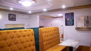 Habitación de hotel con zona de estar que incluye bancos tapizados en amarillo y verde con capitonado, una mesa de mármol y un estante fijado a la pared con adornos.