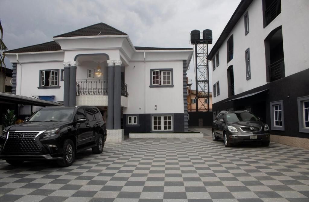 Un bâtiment à deux étages, doté d’une façade blanche et d’un toit noir, avec une allée carrossable à motifs en damier et des voitures garées.