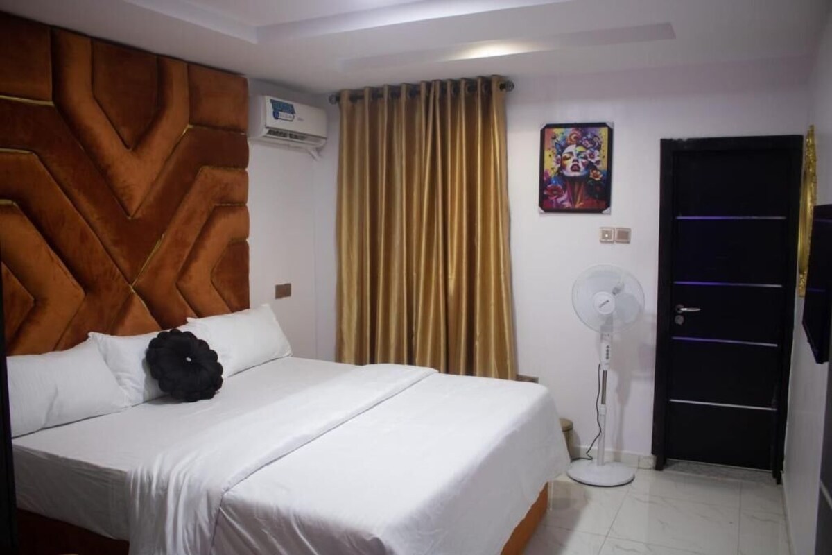 Una camera d'albergo con un letto, una ventola portatile, una porta e un condizionatore d'aria fissato al muro.