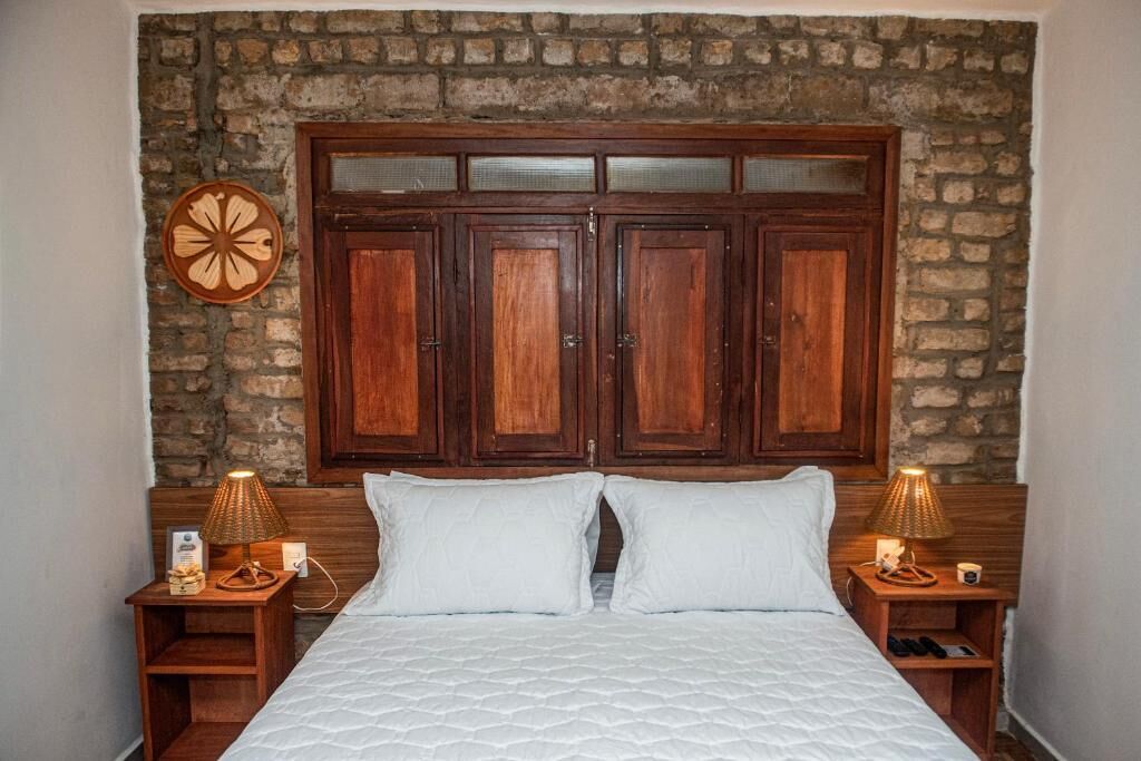 Quarto com grande cabeceira de madeira, cama com roupa de cama branca, duas mesas de cabeceira com abajures e um relógio de parede.