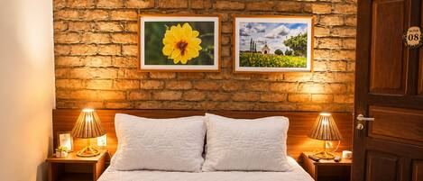 Une chambre d’hôtel avec un mur en briques, deux tableaux encadrés, un lit avec du linge de lit blanc et une porte indiquant le numéro de la suite.