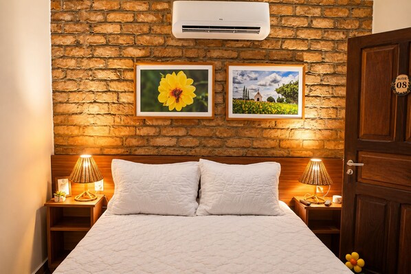 A hotel room with a brick wall, two framed pictures, a bed with white bedding, and a door with a suite number. - A Flor - Pousada (Garanhuns)