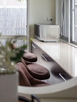 A modern kitchen with a microwave on the counter, a window with a view of a fence, and a plant in the foreground. - Great Value | Private Pool| Walk to Market Square (Sunnybank)
