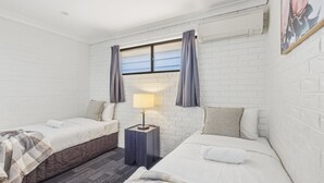 A room with two beds, a nightstand with a lamp, a window with curtains, and a white brick wall. - Affordable townhouse Retreat| Pool| Prime Location (Eight Mile Plains)