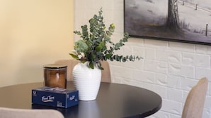 A dining area with a round table, a white vase with green foliage, a box of tissues, and a framed painting of a tree on the wall. - Affordable townhouse Retreat| Pool| Prime Location (Eight Mile Plains)