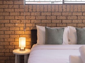 A bed with white bedding and grey pillows, a round bedside table with a lamp, and a brick wall background. - Great value Sunnybank Family Stay | Pool | Parking (Sunnybank)