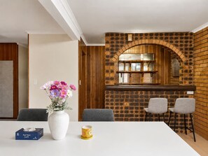 Un interior moderno con una mesa blanca, dos sillas grises, un florero con flores, una caja de pañuelos y una pared de ladrillo con un arco de madera.