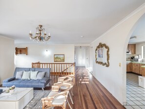 Ein geräumiges Wohnzimmer mit einem blauen Sofa, Korbsesseln, einer Holztreppe und einer durch einen Bogen sichtbaren Küche.