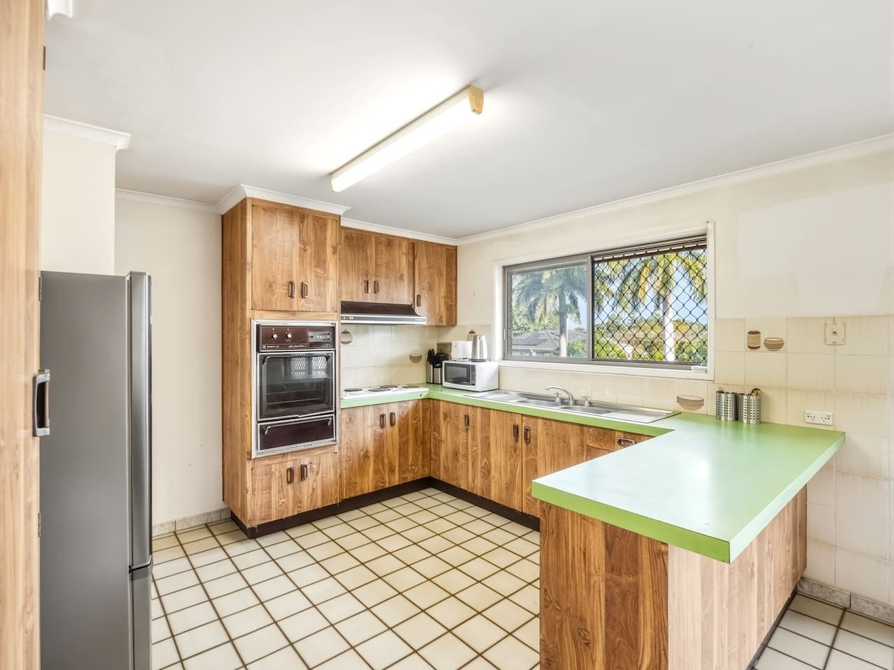 A kitchen with wooden cabinets, a stainless steel refrigerator, a microwave, and a window with a view of greenery.