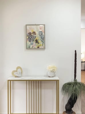 Um console branco com moldura dourada, um vaso branco com flores e uma decoração em formato de coração.