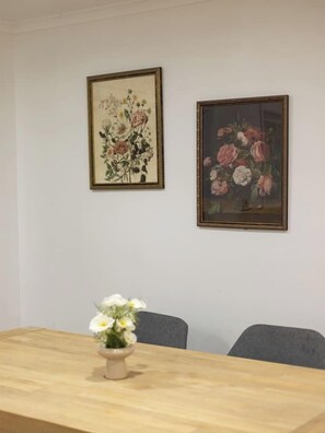 Quarto com mesa de madeira e duas cadeiras, um vaso com flores brancas e dois quadros florais emoldurados na parede.