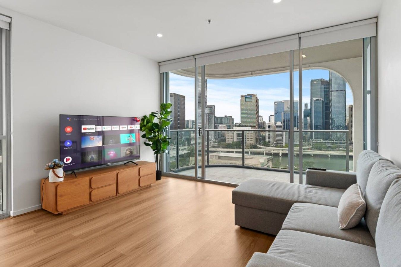 A modern living room with a flat-screen TV, a grey sofa, and a large window offering a city view.