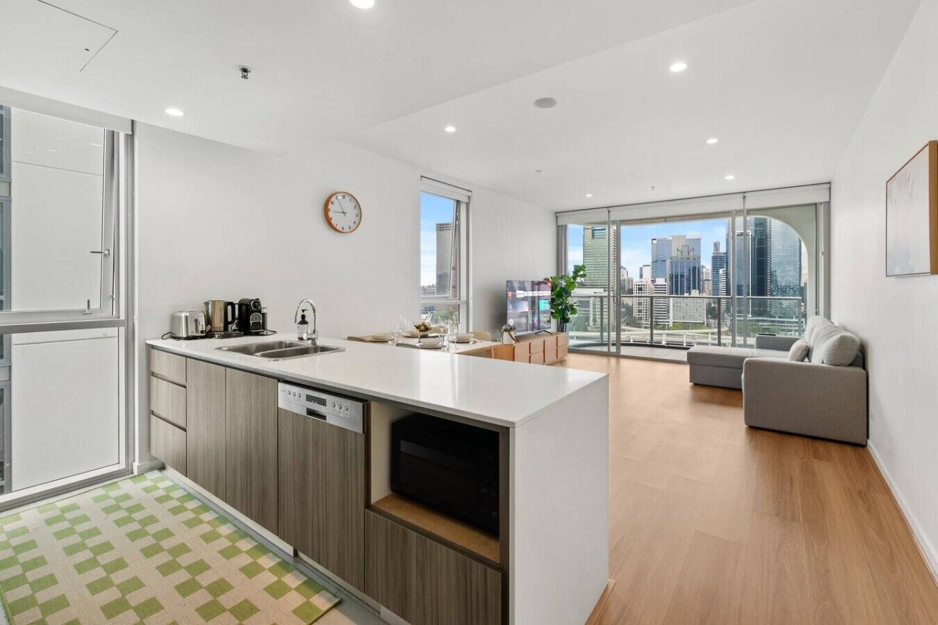 A modern kitchen with a built-in dishwasher, a dining area with a sofa, and a city view through large windows.