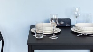 Una mesa de comedor dispuesta con platos, vasos y cubiertos, frente a una pared azul claro con un cuadro abstracto grande encima.