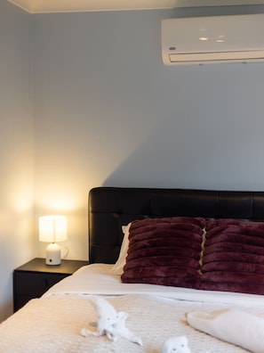 A bedroom with a black headboard, a white lamp on a bedside table, and a large air conditioning unit mounted on the wall.