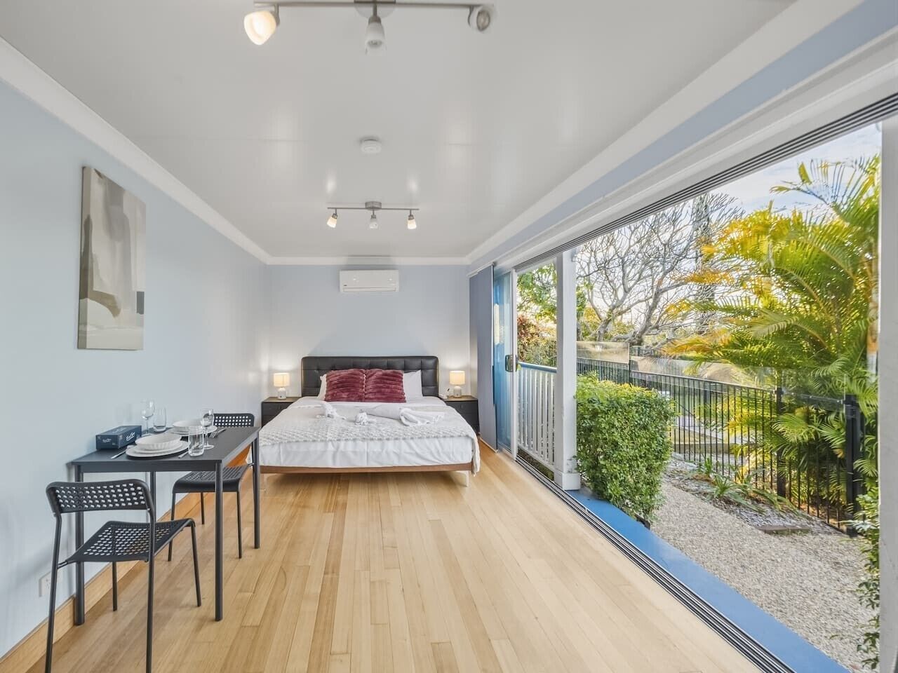 Een slaapkamer met een bed, een bureau en stoelen, een balkon met uitzicht op bomen en een hek.