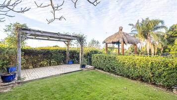 Een tuin met een pergola, een constructie met rieten dak en goed onderhouden hagen.