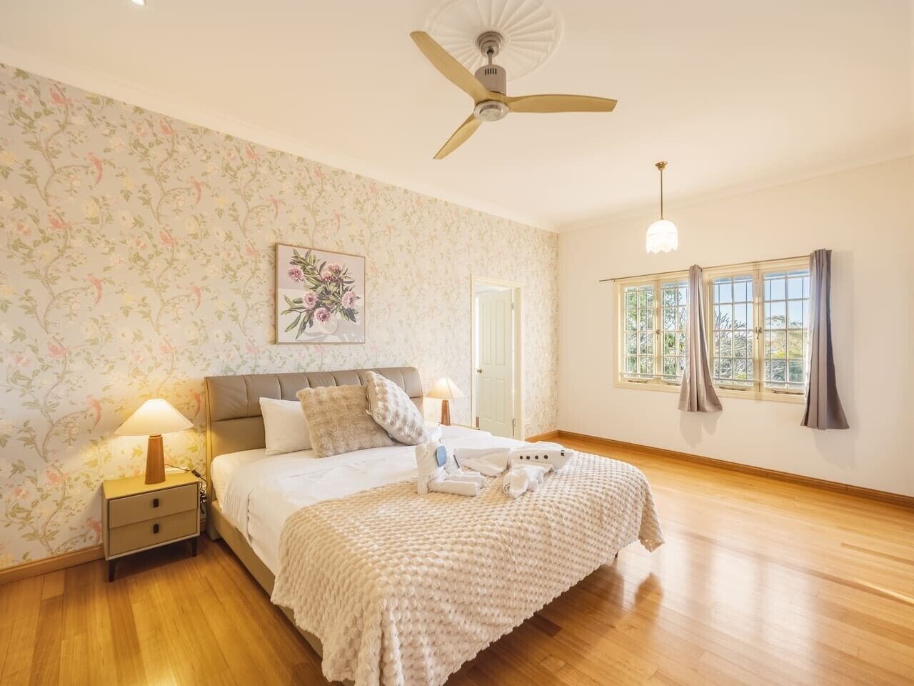 A bedroom with a bed, nightstand, lamp, ceiling fan, window with curtains, and a floral wallpaper.