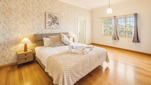 Un dormitorio con cama, mesita de noche, lámpara, ventilador de techo, ventana con cortinas y un papel tapiz floral.