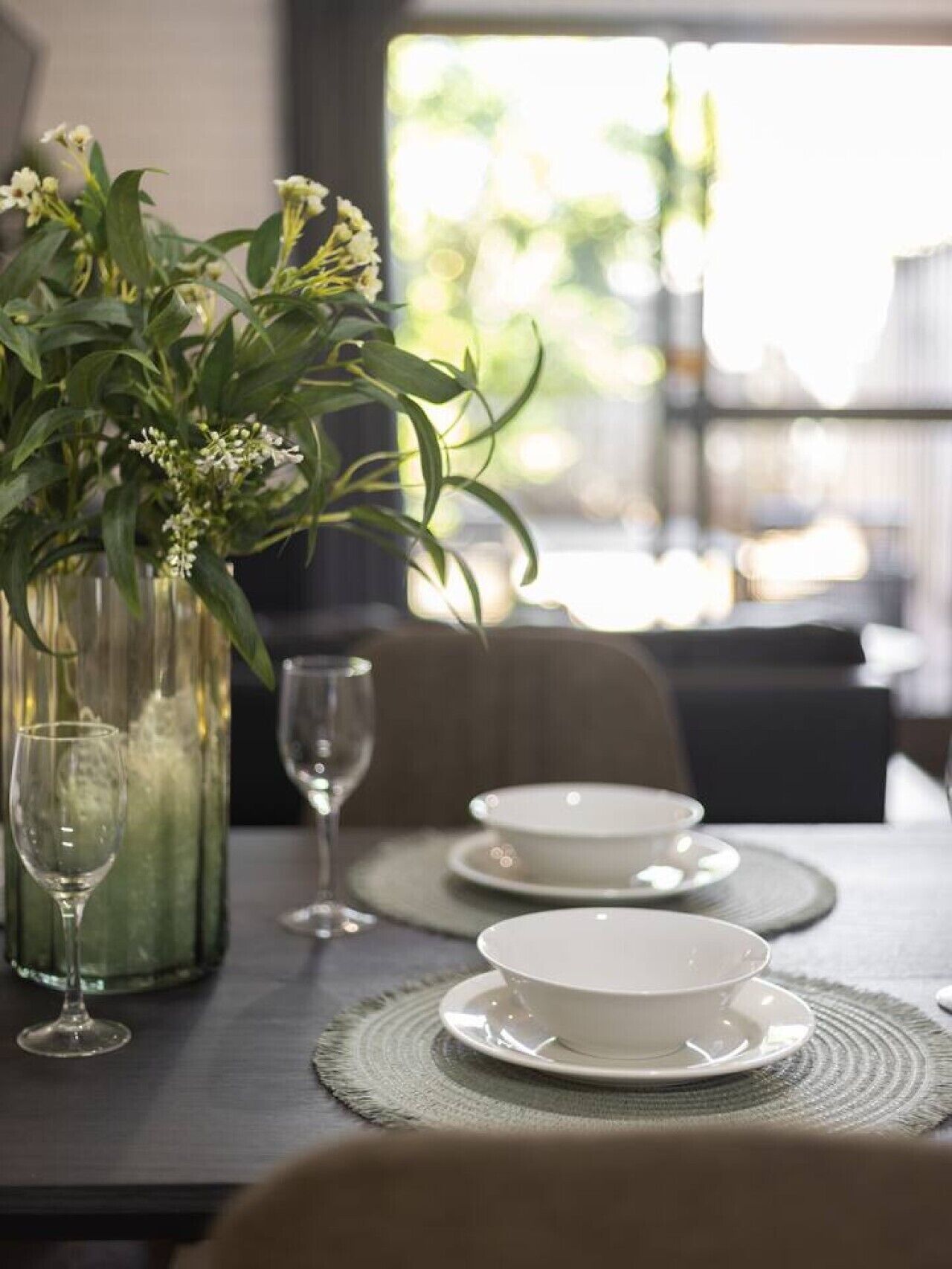 A dining table set with white plates, bowls, and a glass. There is a vase with green foliage and white flowers on the table.