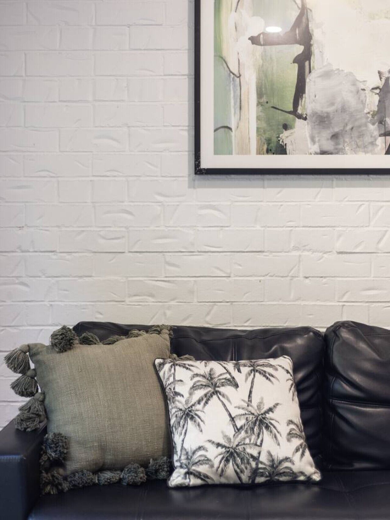 A black leather sofa with two decorative pillows against a white brick wall.