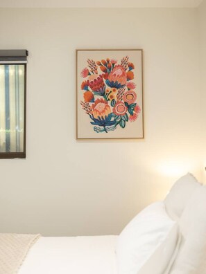 A room with a bed, a window with blinds, and a framed floral artwork on the wall.