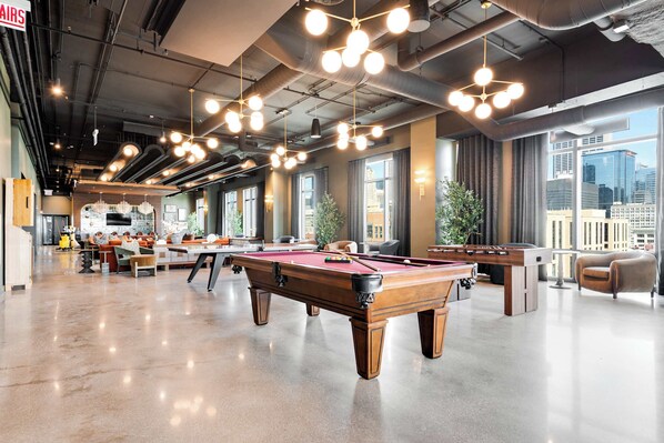 Une salle de billard dotée de plusieurs tables, de grandes fenêtres et d’un plafond haut avec des conduits apparents.