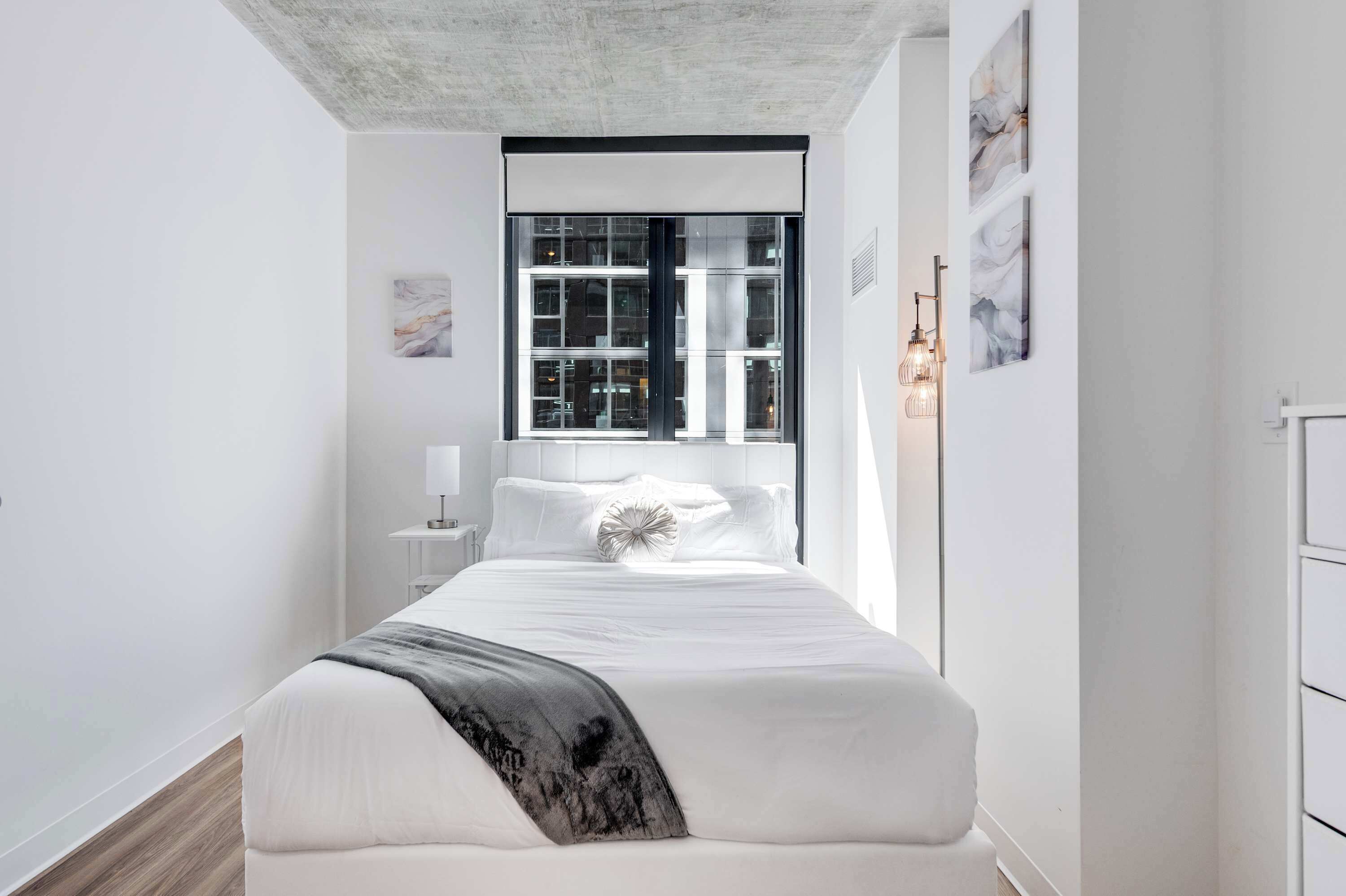 Quarto moderno com uma cama grande, janela com persianas, mesa de cabeceira e abajur.