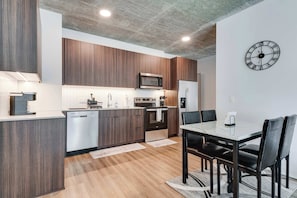 Une cuisine moderne avec des meubles en bois, un four à micro-ondes, un four et un réfrigérateur. On y trouve une table à manger avec des chaises et une horloge au mur.