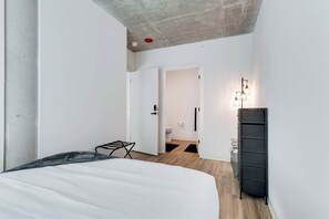 Une chambre à coucher moderne avec un lit, un étendoir à linge en métal noir, une armoire et une salle de bain visible à travers une porte ouverte.