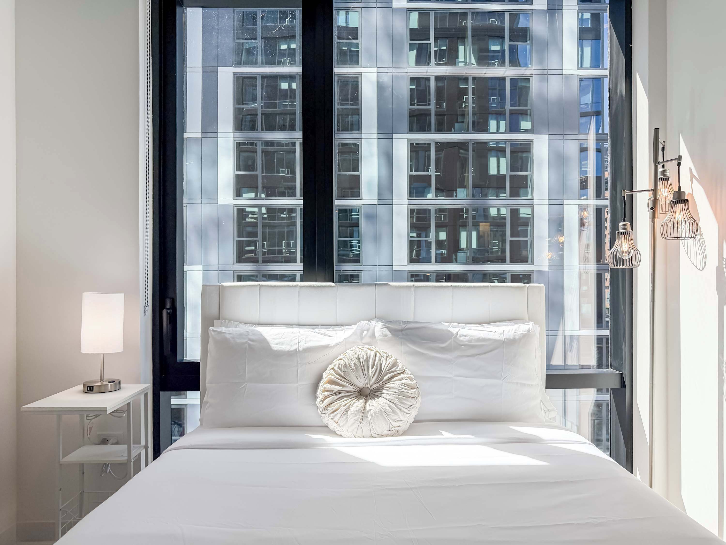 A modern hotel room with a large window, a bed with white linens, a bedside table with a lamp, and a decorative wall light.