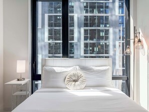 A modern hotel room with a large window, a bed with white linens, a bedside table with a lamp, and a decorative wall light. - Wabash 4BR - Pool, Free In & Out Parking 1 (Chicago)