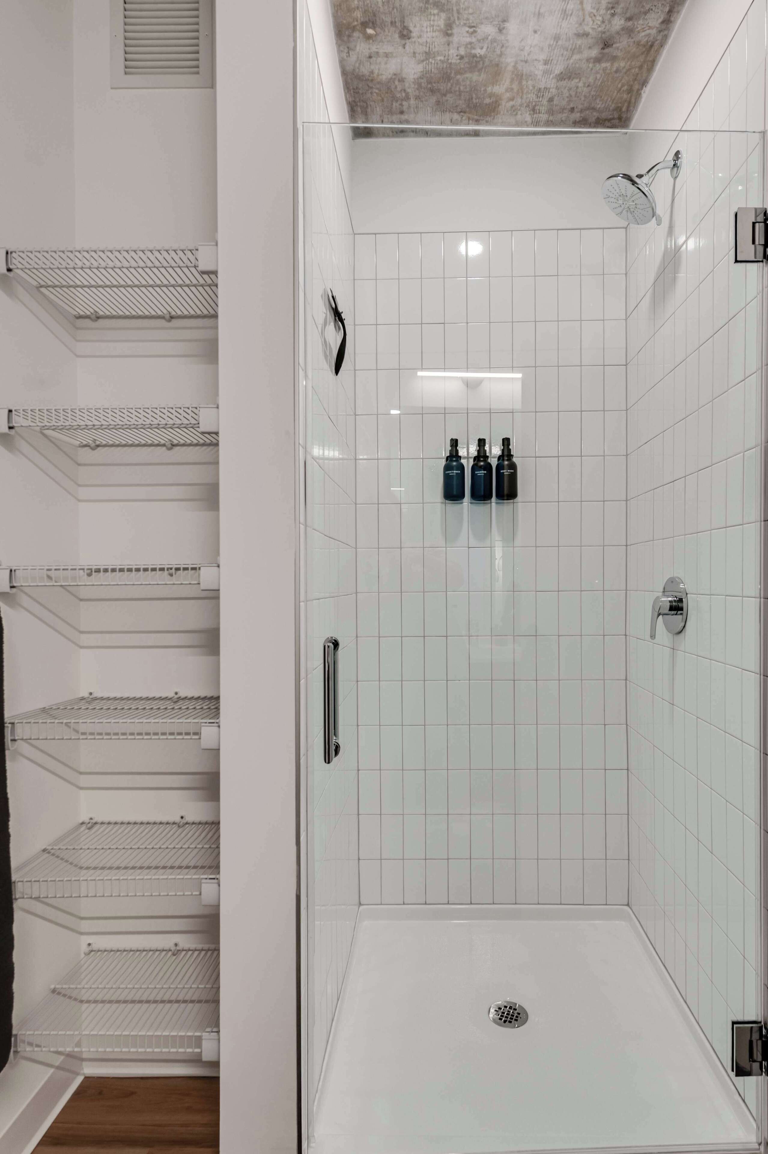 Casa de banho moderna com paredes revestidas de azulejos brancos, cabine de duche em vidro e uma unidade de prateleiras metálicas com espaço de arrumação.