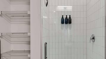 Casa de banho moderna com paredes revestidas de azulejos brancos, cabine de duche em vidro e uma unidade de prateleiras metálicas com espaço de arrumação.