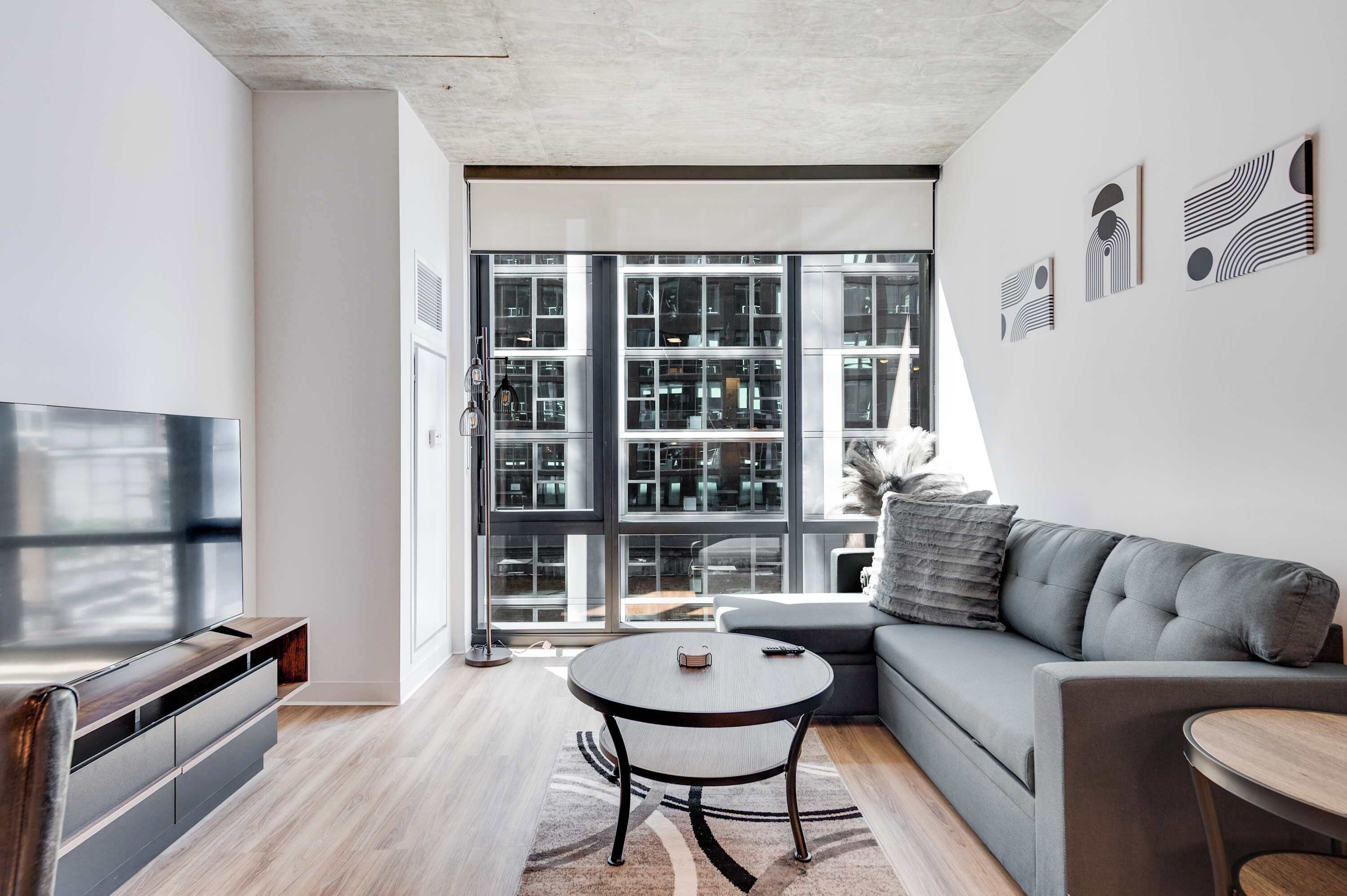 A modern living room with a flat-screen TV, a grey sofa, a round coffee table, and a large window with a view of a building.
