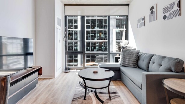 A modern living room with a flat-screen TV, a grey sofa, a round coffee table, and a large window with a view of a building. - Wabash 4BR - Pool, Free In & Out Parking 1 (Chicago)