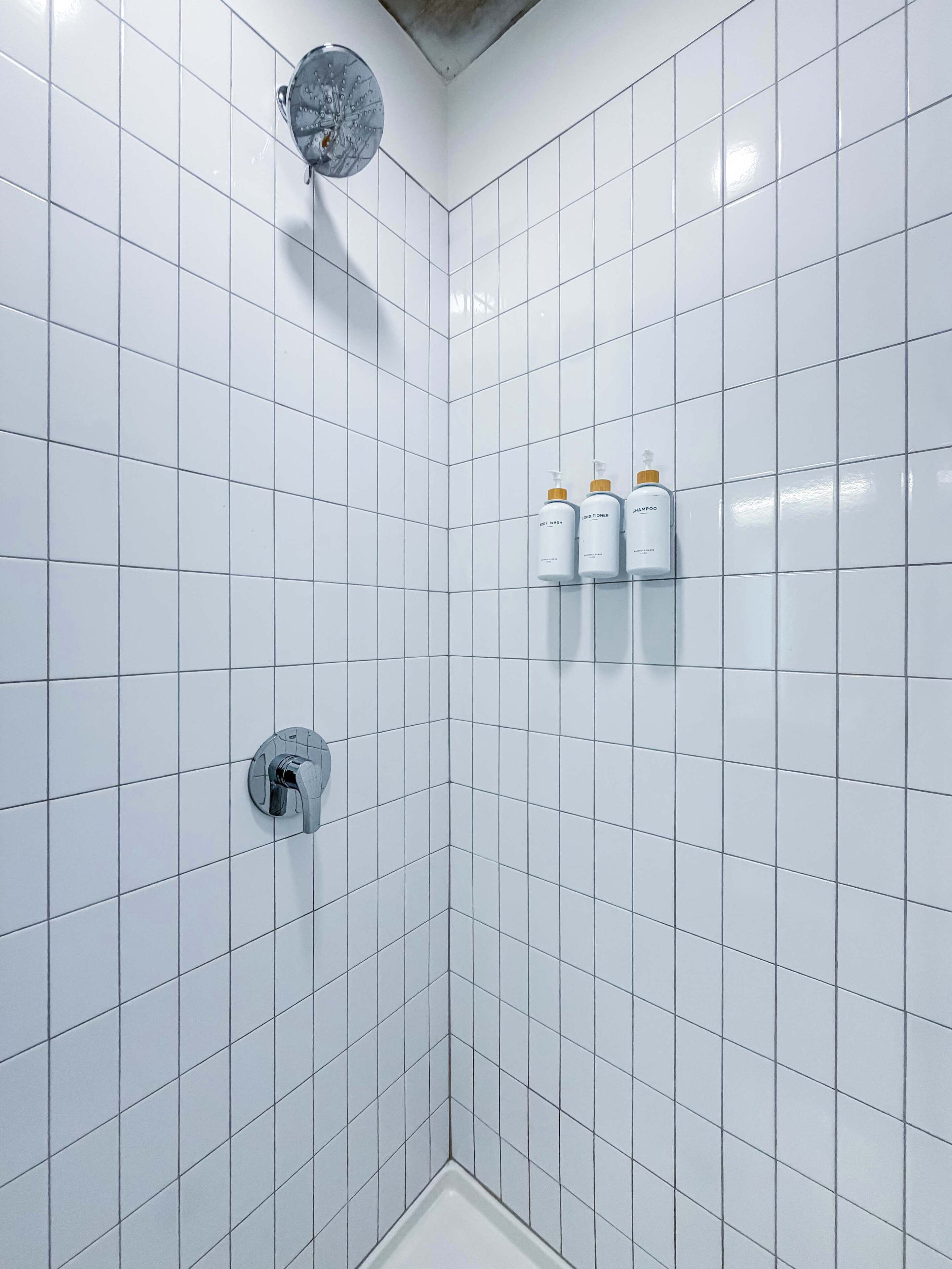 A bathroom with white tiled walls, a showerhead, and three bottles of toiletries.