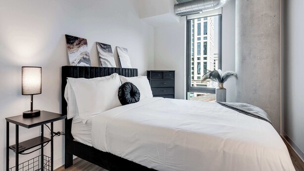 A modern bedroom with a bed, a nightstand, a lamp, and artwork on the wall. - Wabash 4BR - Pool, Free In & Out Parking 2 (Chicago)