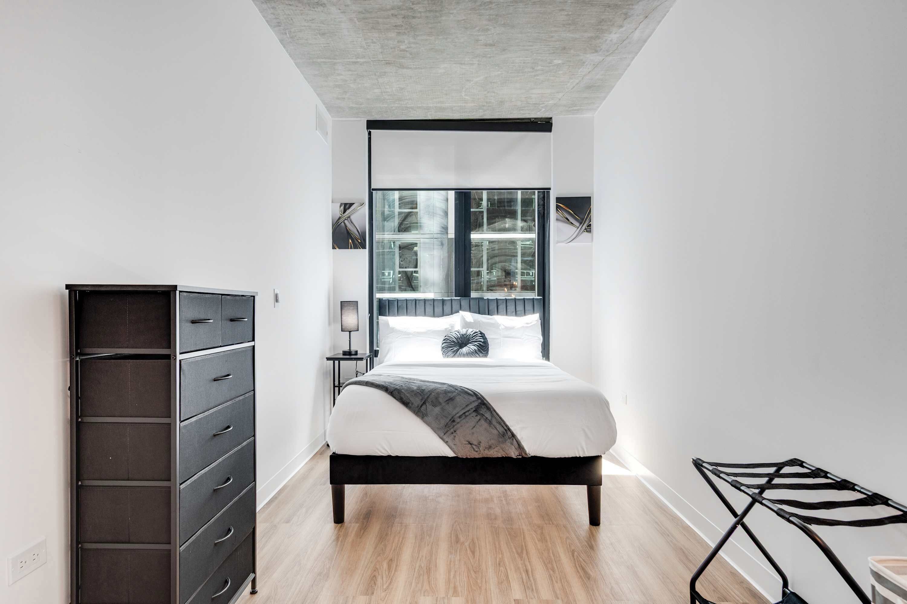 A modern bedroom with a bed, a dresser, a lamp, and a view of the outside.