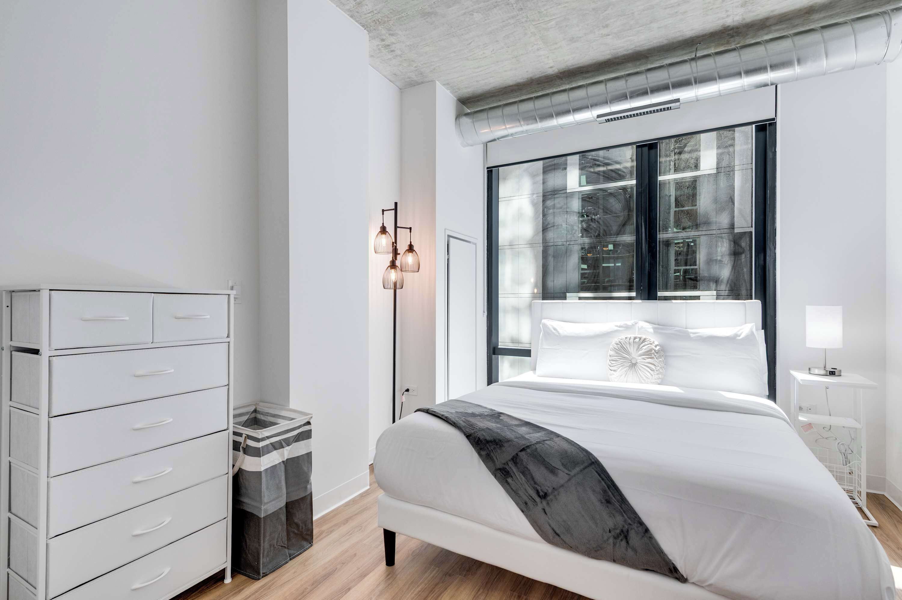 Un dormitorio moderno con una cama grande, un aparador blanco, una lámpara de pie y vistas al exterior a través de un ventanal.